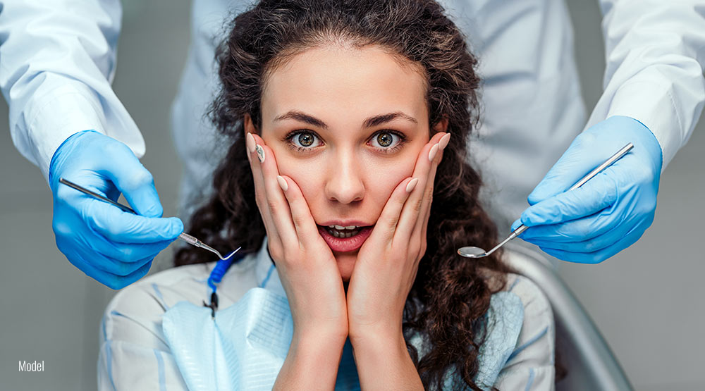 A scared young woman sits in a chair next to a dentist holding dental tools.